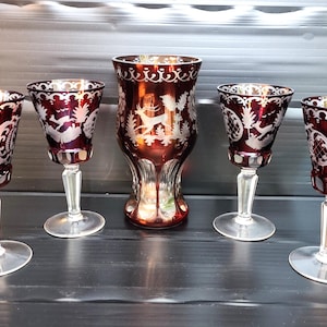 May include: A collection of five antique ruby-coloured glass items. Four stemmed glasses and a vase feature etched white designs. The glasses have a clear stem and base. The vase has a wider base and a detailed etched design.