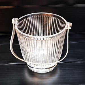 May include: Clear glass ice bucket with a vertical ribbed design. It features a metal handle and a round base. The bucket is empty and rests on a black surface.