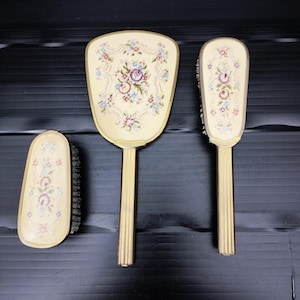 May include: A vintage vanity set featuring a hand mirror, a hairbrush, and a clothes brush. Each piece has a cream-colored background with a floral needlepoint design in pink, blue, and green. The handles and frames are gold-toned.