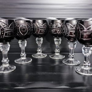 Puede incluir: Seis copas de vino tinto oscuro con intrincados diseños grabados. Las copas tienen tallos y bases transparentes y facetados. El fondo es una superficie oscura y texturizada, creando un contraste.