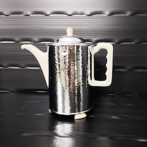 Könnte beinhalten: Eine gehämmerten Edelstahl-Kaffeekanne mit weißem Griff, Ausguss und Deckelknopf. Die Kanne hat einen zylindrischen Körper und einen Deckel. Der Griff ist rechteckig mit einer gebogenen Öffnung. Der Ausguss ist gebogen.