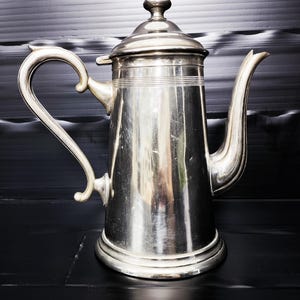 Könnte beinhalten: Eine silberfarbene Kaffeekanne mit einem gebogenen Griff und einem langen, schlanken Ausguss. Die Kanne hat einen Deckel mit einem kleinen Knopf oben und horizontale Linien in der Mitte. Die Kanne ist aus Metall und hat eine glänzende Oberfläche.