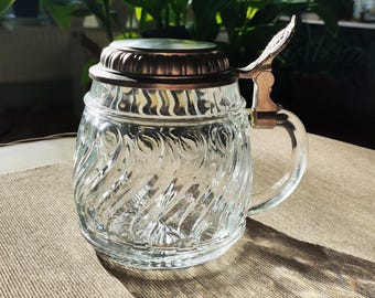 May include: Clear glass beer stein with a decorative swirl pattern. The stein has a metal lid and handle. The lid is a copper colour and the handle is a brass colour. The stein is on a tan surface.