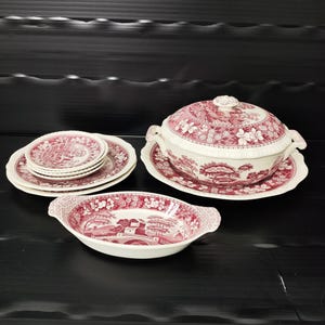 Puede incluir: Una colección de vajilla de cerámica blanca y roja vintage. El juego incluye platos, una fuente para servir y una sopera con tapa. Las piezas presentan un detallado patrón floral y paisajístico. La vajilla está dispuesta sobre una superficie oscura.