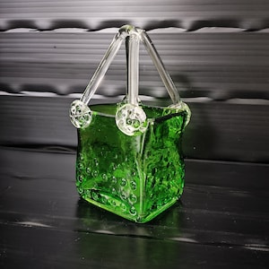 Può includere: Un cestino di vetro verde con un manico in vetro trasparente. Il cestino ha una superficie strutturata con piccole rientranze rotonde. Il manico è attaccato al cestino con sfere decorative di vetro trasparente.