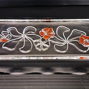 Puede incluir: Una bandeja rectangular de vidrio con un diseño floral. La bandeja presenta una base de vidrio transparente con un patrón decorativo de flores estilizadas en plata y rojo-naranja. La bandeja mide aproximadamente 25 cm de largo y 13 cm de ancho.