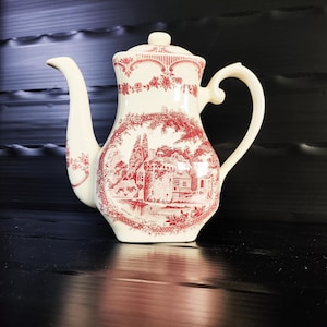 Pode incluir: Cafeteira de cerâmica creme com um design vermelho. O design apresenta uma paisagem com edifícios e árvores. A cafeteira tem um bico curvo, uma alça e uma tampa. Tem aproximadamente 23 cm de altura.