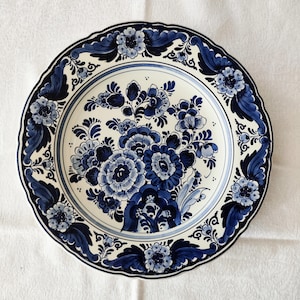 Könnte beinhalten: Ein runder Keramikteller mit gewelltem Rand auf cremefarbenem Grund, verziert mit komplexen dunkelblauen Blumenmustern. Das Design zeigt ein zentrales Blumenbouquet aus stilisierten Blüten und Blättern, umgeben von einer dekorativen Bordüre.