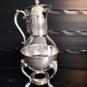 May include: A silver-plated glass coffee server with a decorative handle and lid. The server sits on a silver stand with curved legs and a circular base. The glass carafe has a bulbous shape.