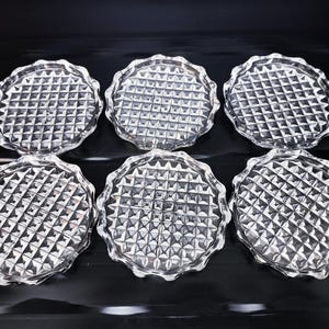 May include: A set of six circular clear glass coasters, each featuring a raised grid pattern of small squares across the surface and a scalloped, wavy edge design. The coasters are arranged in two rows of three, displaying a reflective texture.