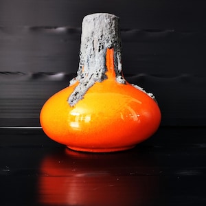 Könnte beinhalten: Eine Keramikvase mit bauchiger Basis und engem Hals. Der untere Teil ist mit einer glatten, glänzenden, leuchtend orangen Glasur versehen, während der obere Hals und Rand eine dicke, strukturierte, poröse graue Lavaglasur aufweisen.
