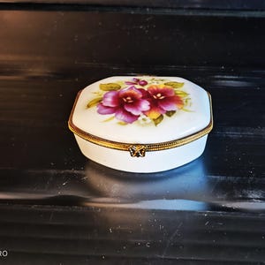 Może przedstawiać: Białe pudełko na drobiazgi z porcelany z pozłacanym wykończeniem i kwiatowym wzorem przedstawiającym fioletowe i żółte bratki. Pudełko ma klapkę z zawiasem z motywem motyla.