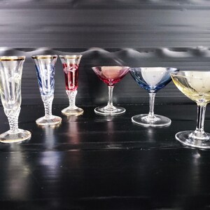 May include: A set of six vintage cordial glasses with gold rims. The glasses have clear, blue, red, and yellow bowls. They are displayed on a dark surface. The glasses are designed with elegant stems and bowls, showcasing a variety of colours.