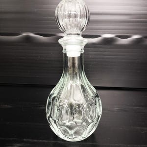 Puede incluir: Una botella de vidrio transparente con un tapón decorativo. La botella tiene una base redondeada con un patrón ovalado y un cuello delgado. El tapón tiene un diseño redondo y acanalado. La botella está vacía y se encuentra sobre una superficie oscura.