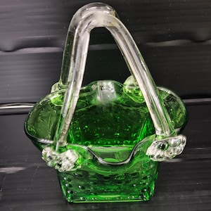 May include: A green glass basket with a clear glass handle. The basket has a square base and a rounded top with a textured surface. The handle is arched and attached to the basket with decorative clear glass accents.
