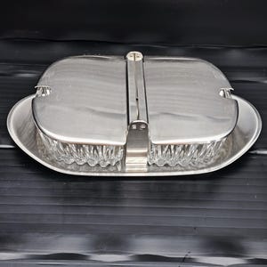 Könnte beinhalten: Eine silberfarbene Servierschale mit Klappdeckel. Die Schale hat eine rechteckige Form mit abgerundeten Kanten und einem transparenten, strukturierten Glaseinsatz. Der Deckel wird mit einem Metallverschluss gesichert. Die Schale steht auf einer schwarzen Oberfläche.