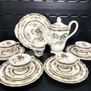 May include: A vintage tea set comprising a white teapot, creamer, serving tray, and teacups with saucers. The set is adorned with floral patterns in blue, purple, and red, with a maroon border. The set is arranged on a black surface.