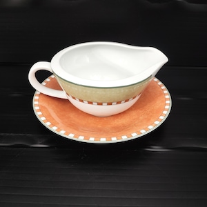 May include: A white ceramic gravy boat with a handle and spout, resting on a matching saucer. The boat and saucer feature a decorative orange and green pattern with a checkered border. The set is designed for serving sauces and gravies.