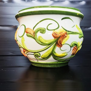 Op de afbeelding: Witte keramische bloempot met een groene rand en basis, versierd met oranje, gele en groene bloemmotieven. De handgeschilderde details creëren een levendig en decoratief stuk, geschikt voor binnen of buiten.
