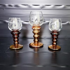 Peut inclure: Trois verres à pied ornés avec des motifs floraux gravés. Les bases et les tiges sont ambrées et marron, avec un design bulbeux et superposé. Les bols en verre clair présentent un motif floral délicat gravé. Les verres sont disposés sur une surface sombre.