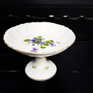 Puede incluir: Un soporte para pasteles de porcelana blanca con borde festoneado y ribete dorado. El soporte presenta un diseño floral de violetas moradas y hojas verdes. La base también está decorada con pequeños detalles florales.