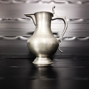 May include: A silver-coloured jug with a curved handle and a spout. The jug has a bulbous body and a lid with a small handle. It is set against a dark background.