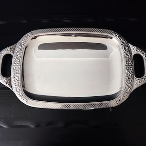 May include: A rectangular silver serving tray with ornate handles and a decorative border. The tray has a reflective surface and is set against a dark background. The handles feature intricate floral designs.