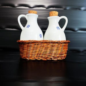 Puede incluir: Dos vinagreras de cerámica blanca con asas y tapones de corcho, cada una con pequeños motivos florales azules, están colocadas juntas dentro de una cesta ovalada de mimbre marrón. Las vinagreras tienen un acabado liso y brillante.