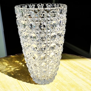 May include: A clear, cut-glass vase with a textured surface and a flared rim. The vase features a diamond-like pattern, reflecting light. The vase is on a light-coloured wooden surface. The vase's height is approximately 25 cm.