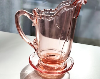 May include: A vintage pink glass creamer with a handle and a decorative rim. The creamer sits on a matching saucer base. The glass has a delicate, translucent quality, and the design features elegant curves and floral accents.