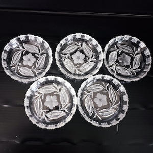 Könnte beinhalten: Fünf klare Glasschalen mit einem Blumen- und Blattdesign. Jede Schale hat einen gewellten Rand und eine Blume in der Mitte, umgeben von Blättern. Die Schalen sind auf einer schwarzen Oberfläche angeordnet.