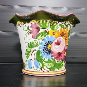 Könnte beinhalten: Ein weißer Keramik-Blumentopf mit einem gewellten grünen Rand und farbenfrohen Blumendesigns. Die handgemalten Blumen umfassen rosa, blaue, gelbe und orangefarbene Blüten mit grünen Blättern und Ranken. Der Topf hat einen gelb-roten Sockel.