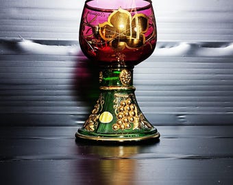 May include: A decorative glass goblet with a red bowl and a green base. The goblet features gold accents and a grape design. The rim of the bowl has a pink hue, and the base is adorned with gold detailing.
