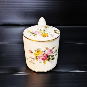 Puede incluir: Jarrón de cerámica blanca con un diseño floral y una tapa. El jarrón tiene un borde dorado y un patrón floral con flores rosas, amarillas y moradas.