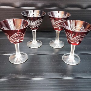 Könnte beinhalten: Vier rubinrote Kristall-Weingläser mit klaren Stielen und Böden. Jedes Glas hat ein geschliffenes Design auf der Schale. Die Gläser sind auf einer dunklen Oberfläche angeordnet.