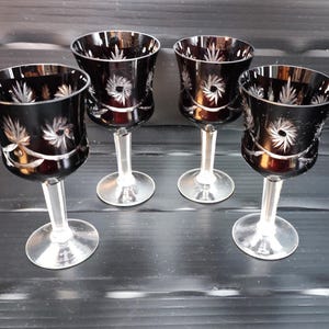 Puede incluir: Cuatro copas de vino tinto oscuro con tallos y bases transparentes. Las copas tienen un diseño grabado de estrellas y hojas. Las copas están dispuestas sobre una superficie oscura.
