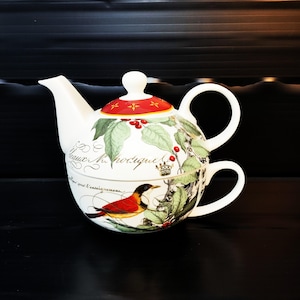 Könnte beinhalten: Weißes Keramik-Teeservice mit rotem und goldenem Deckel. Das Set zeigt ein Vogel- und botanisches Design mit Text. Die Teekanne hat einen gebogenen Ausguss und Griff. Die Tasse ist unter der Teekanne gestapelt.