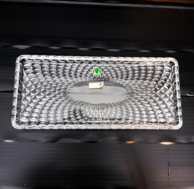 May include: A rectangular clear glass tray with a diamond pattern. The tray has a slightly raised edge and a smooth, oval-shaped center. A small green and white sticker is visible on the tray's surface.