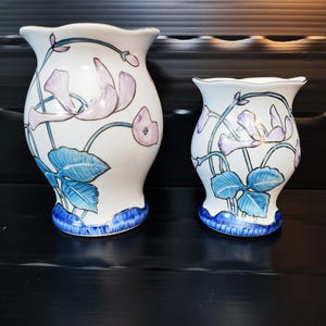 Può includere: Due vasi in ceramica bianca con disegni floreali. I vasi presentano fiori e foglie blu e lavanda. La base di ogni vaso è blu. I vasi sono di diverse dimensioni e poggiano su una superficie scura.