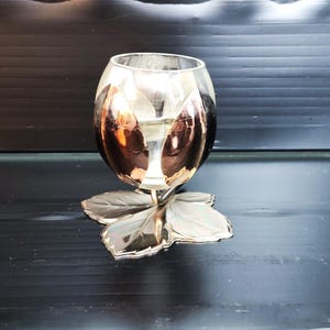 May include: A clear glass candle holder with a copper-coloured design sits on a silver leaf-shaped base.