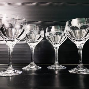 Könnte beinhalten: Vier klare Kristall-Weingläser mit geätzten floralen Designs. Die Gläser haben lange Stiele und breite Kelche, die das Licht reflektieren. Sie sind in einer Reihe angeordnet und zeigen ihre elegante Form und filigranen Details.