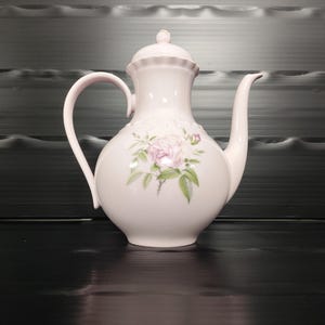 Könnte beinhalten: Weiße Keramik-Teekanne mit Deckel und gebogenem Griff. Die Teekanne hat ein Blumendesign mit rosa Rosen und grünen Blättern. Der Ausguss ist lang und gebogen. Die Teekanne steht auf einer schwarzen Oberfläche.