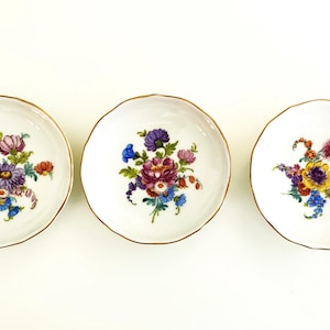 May include: Three small white porcelain dishes with gold trim and floral designs. Each dish features a different arrangement of colorful flowers.