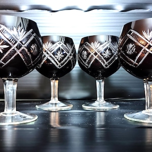 Puede incluir: Cuatro copas de vino de cristal negro con intrincados diseños grabados. Las copas tienen un tallo y una base transparentes, y el cuenco presenta un patrón de estrellas y formas geométricas. Las copas están alineadas.
