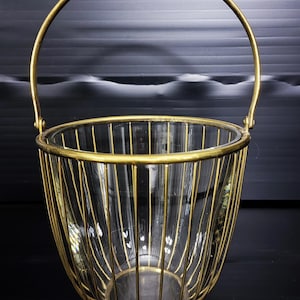 May include: A decorative basket with a clear glass insert and a gold-colored metal cage. The basket has a handle and is designed to hold items. The gold metal cage has vertical bars and a round base.