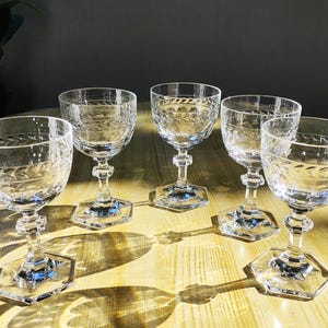May include: Five clear crystal wine glasses with etched floral patterns. Each glass features a hexagonal base and a detailed stem. The glasses are arranged on a light-coloured wooden surface, with shadows cast by the sunlight.
