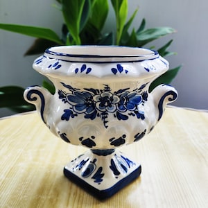 Op de afbeelding: Een witte keramische urnvormige vaas met handgeschilderde blauwe bloemmotieven en krulvormige handvatten. De vaas heeft een brede, geschulpte rand, een afgerond lichaam met reliëfdetails en een vierkante voet met blauwe accenten.