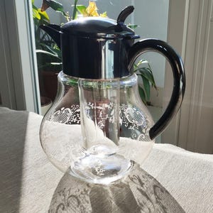 Może przedstawiać: Przezroczysty szklany dzbanek z czarną pokrywką i uchwytem. Dzbanek ma dekoracyjny biały kwiatowy wzór. Dzbanek ma wysokość około 25 cm.