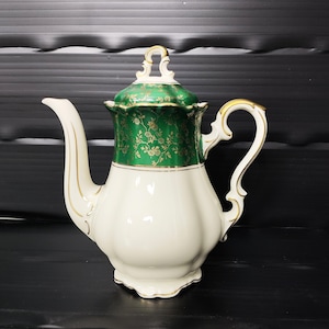 May include: A vintage porcelain teapot. The body is white, and the lid is green with a gold floral pattern. The handle and spout are trimmed with gold. The teapot has a classic design. The height is approximately 20 cm.
