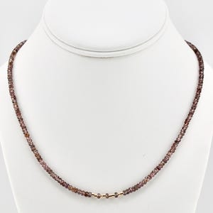 May include: A delicate necklace featuring small, faceted, reddish-brown beads. The beads are strung together, with a few gold-coloured beads near the centre. The necklace is displayed on a white mannequin.